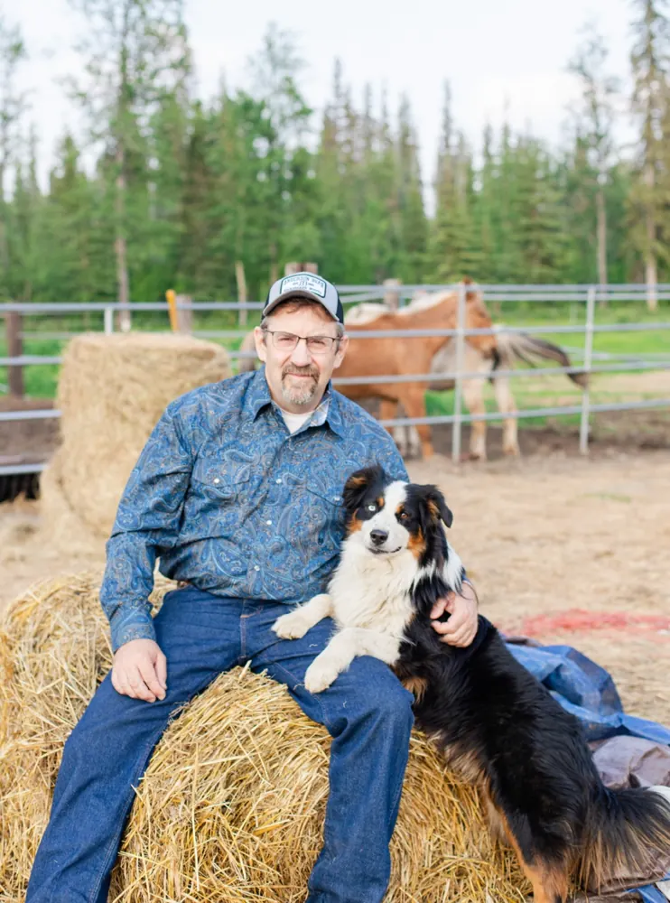 Tom Anderson sitting on a hay bale with a dog, with a horse behind them