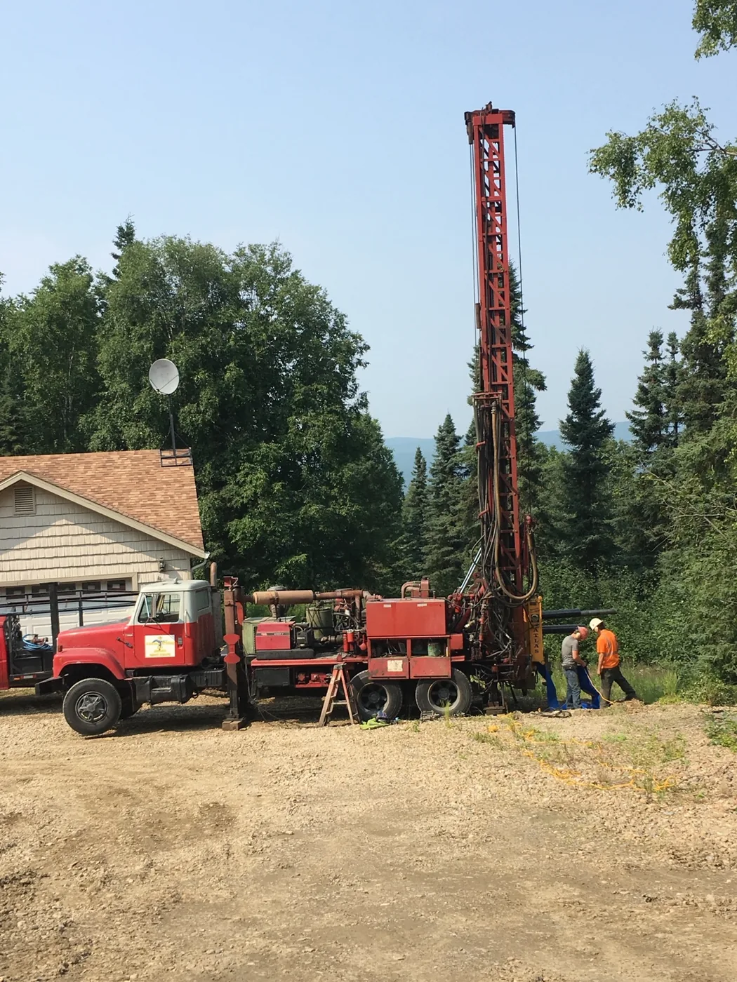 Red water well drilling rig and crew working beside a house